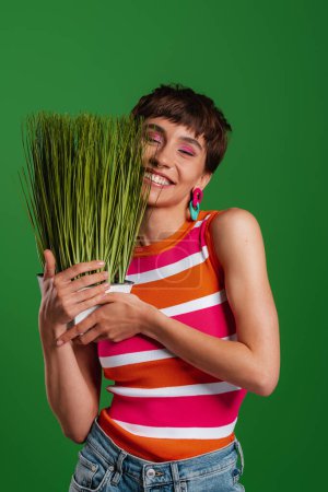 Foto de Hermosa mujer joven sosteniendo planta de interior y sonriendo mientras está de pie sobre fondo verde - Imagen libre de derechos