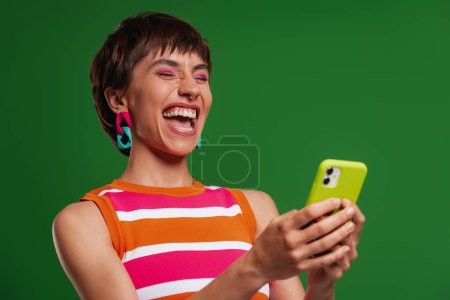 Téléchargez les photos : Jeune femme excitée tenant téléphone intelligent et souriant tout en se tenant debout sur fond vert - en image libre de droit