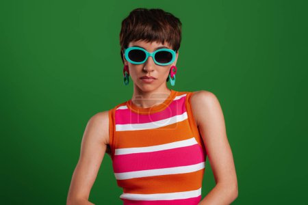 Téléchargez les photos : Jeune femme élégante dans des lunettes à la mode regardant la caméra tout en se tenant debout sur fond vert - en image libre de droit