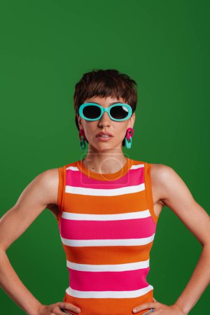 Téléchargez les photos : Jeune femme à la mode dans des lunettes à la mode regardant la caméra tout en se tenant debout sur fond vert - en image libre de droit