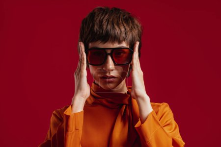 Téléchargez les photos : Portrait de élégante jeune femme aux cheveux courts ajustant ses lunettes à la mode sur fond rouge - en image libre de droit