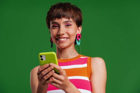 Téléchargez les photos : Belle jeune femme utilisant un téléphone intelligent et souriant tout en se tenant debout sur fond vert - en image libre de droit