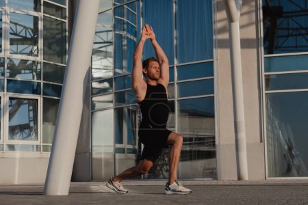 Téléchargez les photos : Athlète masculin confiant faisant des exercices d'étirement à l'extérieur contre le bâtiment urbain - en image libre de droit