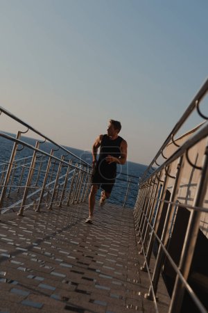 Téléchargez les photos : Beau jeune homme en vêtements de sport profitant du jogging tôt le matin avec la mer sur le fond - en image libre de droit