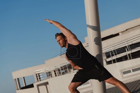 Téléchargez les photos : Homme musclé confiant dans les vêtements de sport faire des exercices d'étirement à l'extérieur tôt le matin - en image libre de droit
