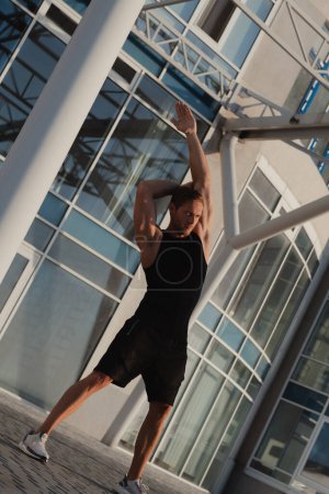 Foto de Hombre musculoso guapo en ropa deportiva haciendo ejercicios de estiramiento al aire libre temprano en la mañana - Imagen libre de derechos