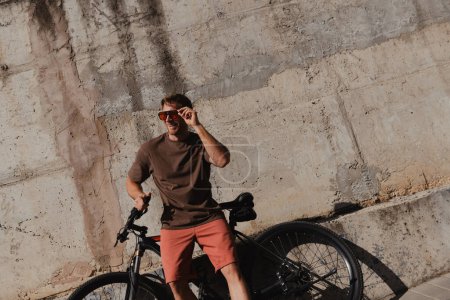 Photo for Confident young man leaning at his bicycle and smiling against a concrete wall outdoors - Royalty Free Image