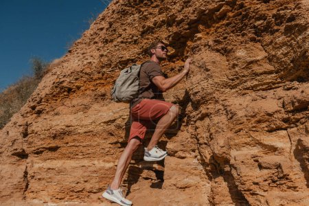 Téléchargez les photos : Homme courageux aventurier avec sac à dos tenant sur la poignée tout en escaladant le rocher - en image libre de droit