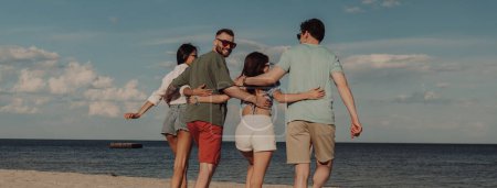 Téléchargez les photos : Vue arrière de jeunes gens joyeux marchant vers la mer au bord de la plage - en image libre de droit