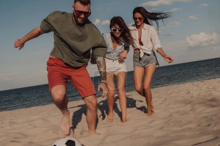 Téléchargez les photos : Groupe de jeunes gens regardant heureux tout en jouant au football sur la plage ensemble - en image libre de droit