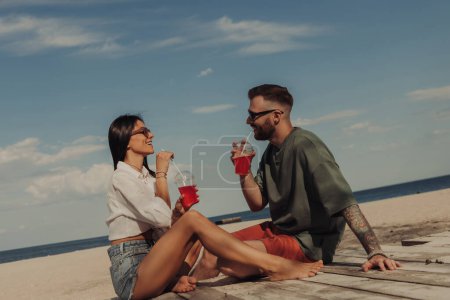 Téléchargez les photos : Joyeux jeune couple profitant de cocktails rafraîchissants tout en se relaxant sur la plage ensemble - en image libre de droit