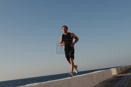 Téléchargez les photos : Jeune homme confiant en vêtements de sport profitant du jogging matinal avec la mer en arrière-plan - en image libre de droit