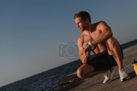 Téléchargez les photos : Athlète masculin confiant vérifiant le programme de remise en forme sur sa montre intelligente tout en s'entraînant bord de mer - en image libre de droit