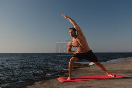 Téléchargez les photos : Confiant jeune homme torse nu faisant des exercices d'étirement à l'extérieur avec la mer sur le fond - en image libre de droit