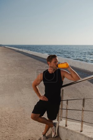 Téléchargez les photos : Vue du dessus de l'eau potable de l'athlète masculin fatigué et vue sur la mer après l'entraînement sportif matinal en plein air - en image libre de droit