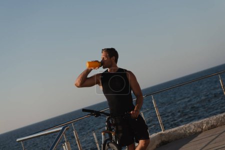 Téléchargez les photos : Jeune homme confiant qui boit de l'eau et se penche sur son vélo avec la mer en arrière-plan - en image libre de droit