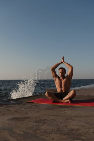 Photo for Handsome young shirtless man sitting on exercise mat while meditating near the sea - Royalty Free Image