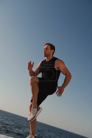 Téléchargez les photos : Jeune athlète masculin confiant en vêtements de sport s'exerçant à l'extérieur avec la mer en arrière-plan - en image libre de droit