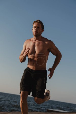 Téléchargez les photos : Confiant jeune homme torse nu jogging à l'extérieur avec la mer sur fond - en image libre de droit