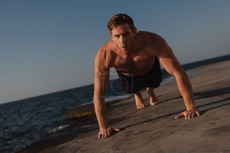 Foto de Joven hombre sin camisa confiado haciendo flexiones mientras hace ejercicio al aire libre con el mar en el fondo - Imagen libre de derechos