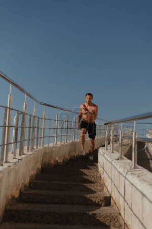 Téléchargez les photos : Athlète masculin confiant descendant les escaliers à l'extérieur avec un ciel bleu en arrière-plan - en image libre de droit