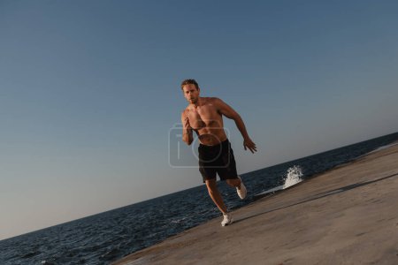 Foto de Hombre atlético seguro con torso muscular corriendo al aire libre con el mar en el fondo - Imagen libre de derechos