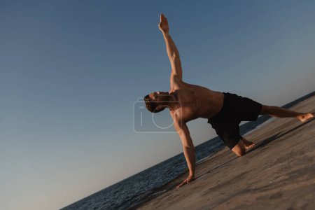 Téléchargez les photos : Beau jeune homme torse nu pratiquant le yoga en plein air avec la mer sur fond - en image libre de droit