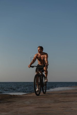 Téléchargez les photos : Beau jeune homme torse nu profitant du matin d'été tout en faisant du vélo bord de mer - en image libre de droit