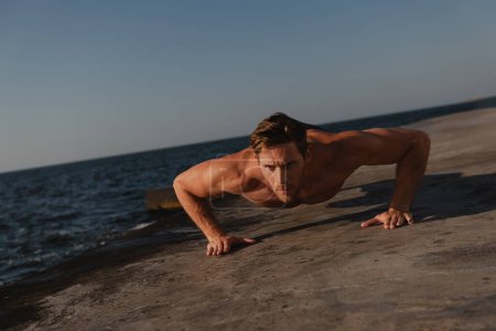 Foto de Guapo atleta masculino haciendo flexiones mientras hace ejercicio al aire libre con el mar en el fondo - Imagen libre de derechos