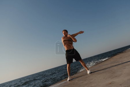 Foto de Longitud completa del hombre musculoso haciendo ejercicios de estiramiento al aire libre con el mar en el fondo - Imagen libre de derechos