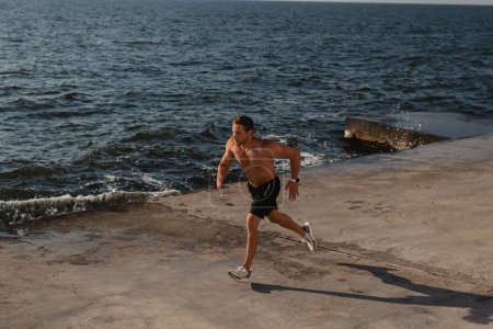 Téléchargez les photos : Vue du dessus de l'homme athlétique confiant jogging par la jetée avec la mer en arrière-plan - en image libre de droit
