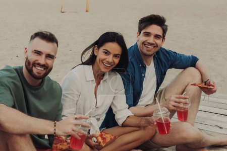 Foto de Jóvenes felices disfrutando de comida y bebidas mientras pasan tiempo juntos en la playa - Imagen libre de derechos