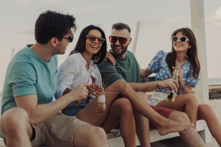Téléchargez les photos : Groupe de jeunes gens parlant et souriant tout en appréciant la bière fraîche sur la plage ensemble - en image libre de droit
