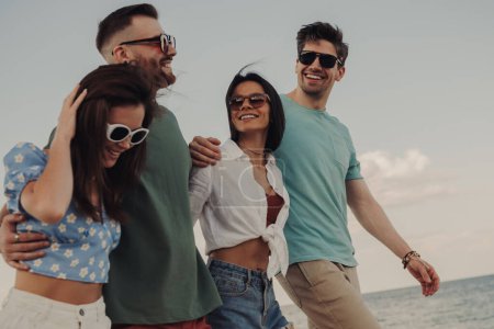 Téléchargez les photos : Groupe de jeunes gens joyeux collant et souriant tout en marchant sur la plage ensemble - en image libre de droit