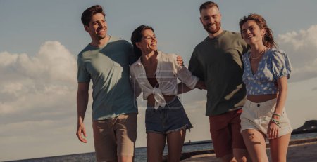 Téléchargez les photos : Groupe de jeunes amis joyeux collant et souriant tout en marchant sur la plage ensemble - en image libre de droit
