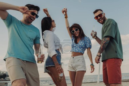 Téléchargez les photos : Groupe de jeunes gens joyeux dégustant de la bière fraîche tout en dansant sur la plage ensemble - en image libre de droit