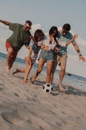 Téléchargez les photos : Quatre jeunes gens heureux passent du temps sans soucis ensemble tout en jouant au football sur la plage - en image libre de droit