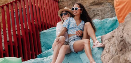 Téléchargez les photos : Deux belles jeunes femmes embrassant et souriant tout en étant assis sur des rochers colorés ensemble - en image libre de droit