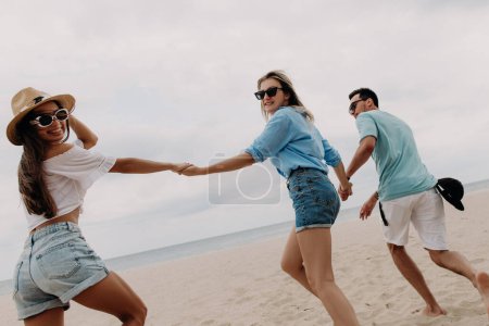 Foto de Tres alegres jóvenes amigos pasando tiempo divertido mientras corren por la playa hacia el mar juntos - Imagen libre de derechos