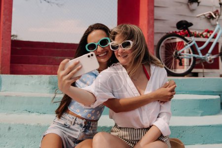 Foto de Dos mujeres jóvenes atractivas haciendo selfie y sonriendo mientras están sentadas al aire libre juntas - Imagen libre de derechos