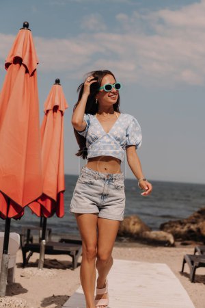 Téléchargez les photos : Jolie jeune femme ajustant les cheveux et souriant tout en profitant de la journée d'été sur la plage - en image libre de droit