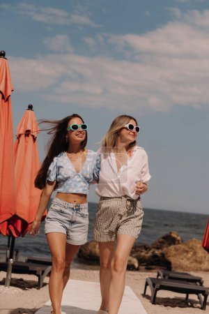 Téléchargez les photos : Deux jeunes femmes joyeuses embrassant et souriant tout en marchant sur la plage ensemble - en image libre de droit