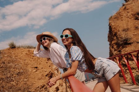 Téléchargez les photos : Deux jeunes femmes à la mode dans des lunettes à la mode souriant tout en passant la journée d'été au bord de la mer ensemble - en image libre de droit