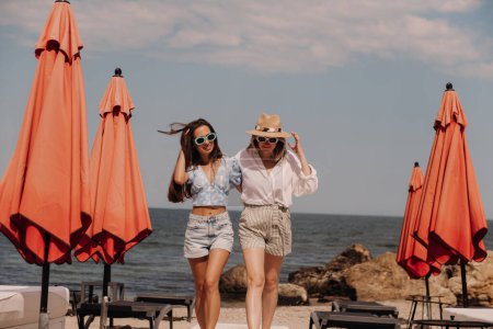 Téléchargez les photos : Deux belles jeunes femmes embrassant et souriant tout en marchant sur la plage ensemble - en image libre de droit