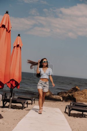 Téléchargez les photos : Pleine longueur de jeune femme heureuse profitant de la journée d'été tout en marchant sur la plage - en image libre de droit