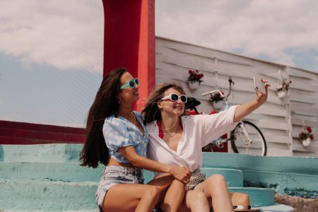 Téléchargez les photos : Deux jolies jeunes femmes faisant du selfie et souriant tout en étant assis à l'extérieur ensemble - en image libre de droit