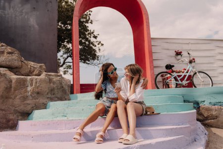 Téléchargez les photos : Deux jeunes femmes heureuses utilisant un téléphone intelligent tout en étant assis à l'extérieur ensemble - en image libre de droit
