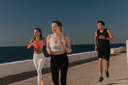 Téléchargez les photos : Trois jeunes gens confiants en vêtements de sport profitant du jogging matinal en bord de mer ensemble - en image libre de droit