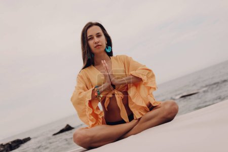 Photo for Beautiful young woman in boho styled crop top looking at camera while meditating on the beach - Royalty Free Image
