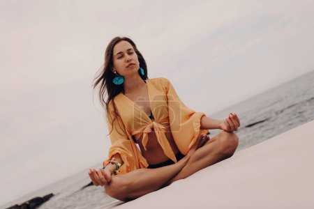 Photo for Beautiful young woman in boho styled crop top sitting in lotus position while meditating seaside - Royalty Free Image